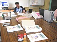 講習会に使うテキストを製本しているところ。画面の奥では、村上さんが湯飲みにストローを差してお茶を飲んでいます。